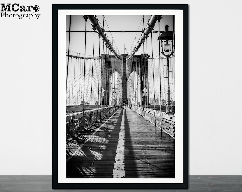 Peut inclure: Une photographie en noir et blanc du pont de Brooklyn &agrave; New York. La photo montre la passerelle du pont avec une vue sur les c&acirc;bles et les tours du pont.