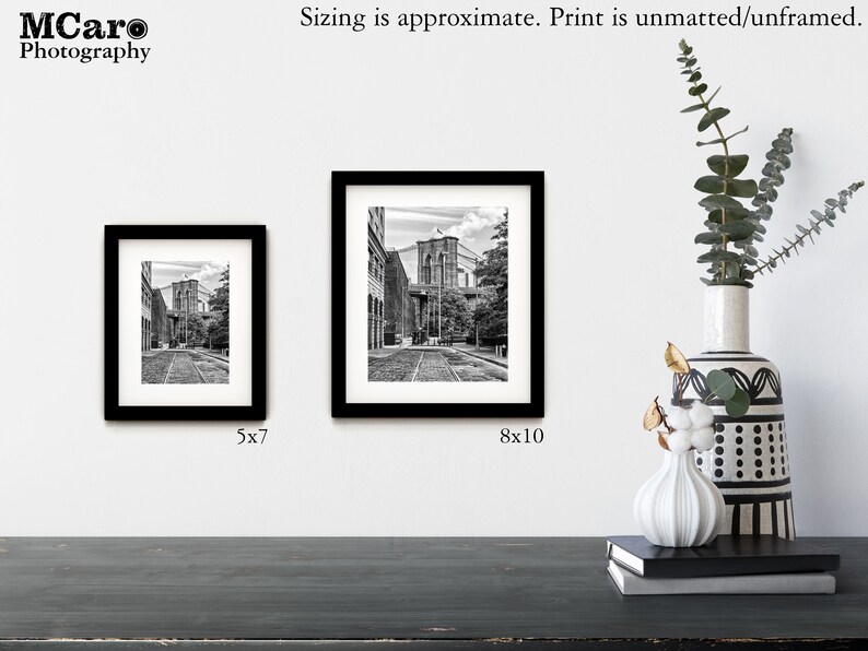 May include: Two black framed photographs of a black and white image of a street in New York City with the Brooklyn Bridge in the background. The smaller photo is 5x7 inches and the larger photo is 8x10 inches.