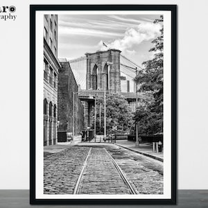 May include: A black and white photograph of the Brooklyn Bridge in New York City. The bridge is seen from a street level perspective, with the bridge's towers and cables in the background. The street is cobblestone and there are buildings on either side of the street.