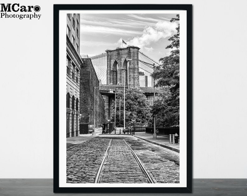 May include: A black and white photograph of the Brooklyn Bridge in New York City. The bridge is seen from a street level perspective, with the bridge's towers and cables in the background. The street is cobblestone and there are buildings on either side of the street.