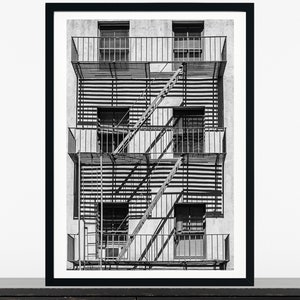 May include: Black and white photograph of a fire escape on the side of a building. The fire escape has four levels and is made of metal. The building is made of brick and has windows on each level.