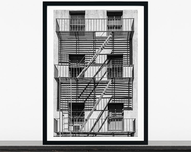 May include: Black and white photograph of a fire escape on the side of a building. The fire escape has four levels and is made of metal. The building is made of brick and has windows on each level.