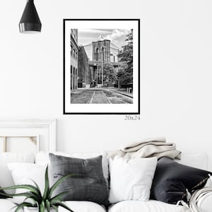 May include: Black and white photograph of the Brooklyn Bridge in New York City. The bridge is seen from a street level perspective with buildings and trees in the foreground. The image is framed in a black frame and measures 20x24 inches.