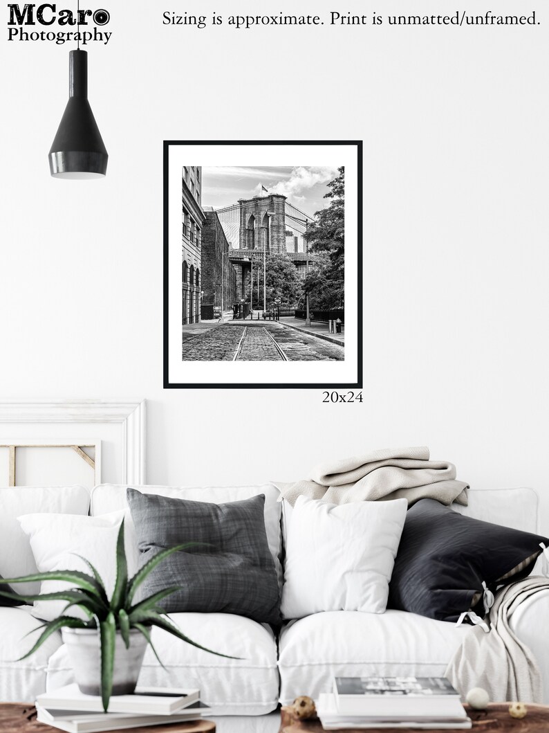May include: Black and white photograph of the Brooklyn Bridge in New York City. The bridge is seen from a street level perspective with buildings and trees in the foreground. The image is framed in a black frame and measures 20x24 inches.