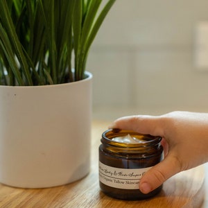 May include: A brown glass jar with a white label that reads "Farm Baby & Kids Super Cream Organic Tallow Skincare". The jar is filled with a white cream and is being held by a hand.