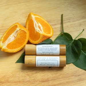 May include: Two wooden lip balm tubes with white labels. The top label reads "INGREDIENTS: grass fed tallow, beeswax, olive oil, jojoba oil, castor oil, emu oil, essential oils of eucalyptus & wild orange, vitamin e oil" and the bottom label reads "SEFORA'S ORGANIC TALLOW SKINCARE EUCALYPTUS WILD ORANGE". The tubes are sitting on a wooden surface with two orange halves and green leaves.