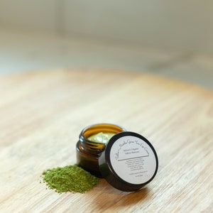 May include: A small glass jar with a green lid containing a green cream. The jar is open and the lid is sitting next to it. There is a small pile of green powder next to the jar. The label on the lid reads "Sulfur's Organic Tallow Skincare Matcha Green Tea Eye Cream".