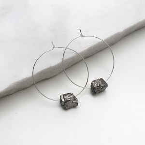 Peut inclure: Une paire de boucles d'oreilles créoles argentées avec une pierre cubique gris foncé attachée à chaque créole.