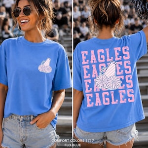 Op de afbeelding: Een blauw t-shirt met een kleine afbeelding op de linkerborst van een megafoon en pompons. De achterkant toont het woord EAGLES in grote roze blokletters die drie keer worden herhaald, gecentreerd met een afbeelding van een megafoon en pompons.