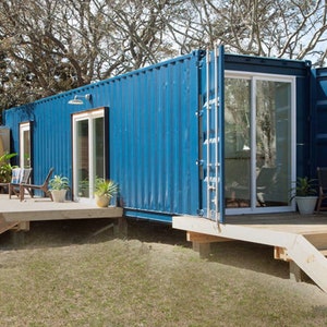 May include: A blue shipping container home with a wooden deck and stairs. The container has large glass doors and windows, with a surfboard leaning against a wooden privacy screen. Potted plants and outdoor furniture are visible.
