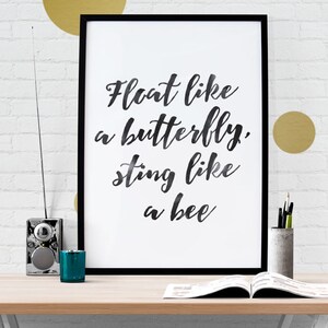 May include: A framed print with the quote "Float like a butterfly, sting like a bee" in black script. The print is on a desk with a radio, a teal candle holder, a concrete pencil holder, and an open book. Gold circles decorate the white brick wall.