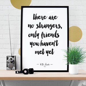 May include: A black framed print displaying the quote "there are no strangers, only friends you haven't met yet" in black script. A small radio, headphones, and a potted plant are on a wooden surface.