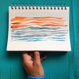 Op de afbeelding: Een aquarel schilderij van een zonsondergang boven de oceaan. Het schilderij is op een wit vel papier met een spiraalbinding. Het schilderij heeft oranje en rode strepen aan de bovenkant, die de zonsondergang voorstellen, en blauwe en grijze strepen aan de onderkant, die de oceaan voorstellen.