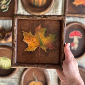 Könnte beinhalten: Ein quadratisches, gerahmtes Kunstwerk mit einer detaillierten Malerei eines Herbstblattes in Gelb-, Orange- und Grüntönen. Das Kunstwerk wird in einer Hand gehalten, im Hintergrund sind weitere runde und quadratische Kunstwerke mit Herbstmotiven zu sehen.