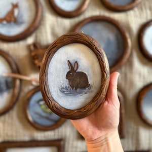May include: A circular painting of a brown rabbit, set within a wooden frame with a textured, bark-like design. Other framed animal paintings are visible in the background, creating a collection.