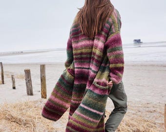 May include: A long-sleeved, knee-length cardigan featuring a horizontal striped pattern in shades of magenta, olive green, and dark teal. The knit garment has a relaxed fit, long sleeves, and visible side pockets, worn over plain olive green trousers.