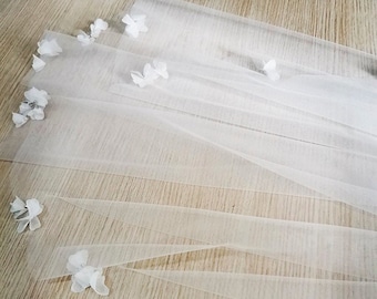 May include: A sheer, ivory bridal veil adorned with delicate, white floral embellishments. The veil is layered and features the text "MI BRIDAL VEIL". The flowers are clustered and scattered across the fabric, adding a touch of elegance.