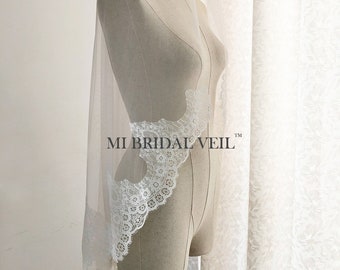 May include: A sheer white bridal veil with a delicate lace trim along the edge. The veil is draped over a mannequin, showcasing its flowing design. The text "MI BRIDAL VEIL" is printed on the veil.