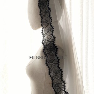 May include: A bridal veil with a delicate black lace trim. The veil is sheer and white, with the lace adding a contrasting dark accent. The lace design is intricate, featuring floral patterns. The text "MI BRIDE VEIL" is visible on the mannequin.