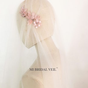 May include: A bridal veil with a floral headpiece. The headpiece features pink fabric flowers and leaves with sparkling embellishments. The veil is a sheer, light pink color. The text "MI BRIDAL VEIL" is visible.