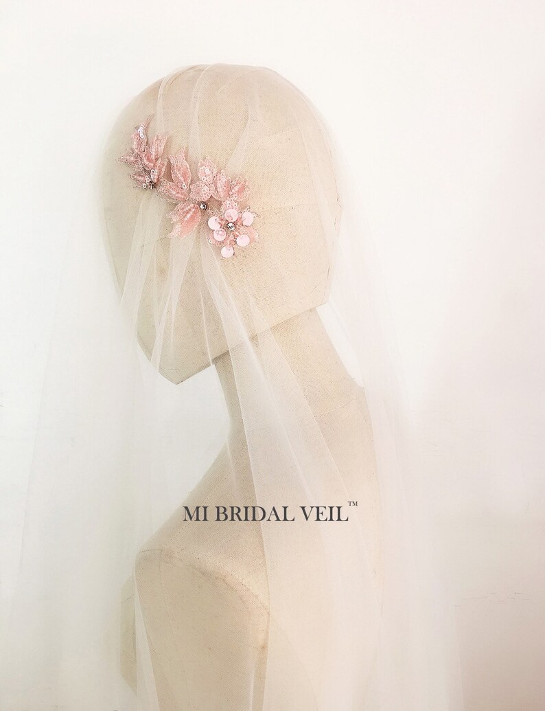 May include: A bridal veil with a floral headpiece. The headpiece features pink fabric flowers and leaves with sparkling embellishments. The veil is a sheer, light pink color. The text "MI BRIDAL VEIL" is visible.