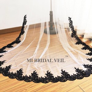 May include: A white bridal veil with a black floral lace trim. The veil is displayed on a stand, showcasing its length and delicate design. The text "MI BRIDAL VEIL" is visible on the veil.
