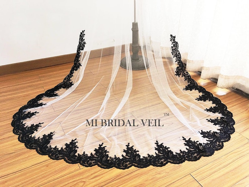 May include: A white bridal veil with a black floral lace trim. The veil is displayed on a stand, showcasing its length and delicate design. The text "MI BRIDAL VEIL" is visible on the veil.