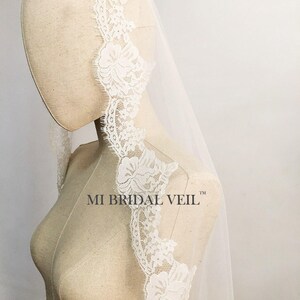 May include: A white bridal veil with a delicate floral lace trim. The lace is intricately designed with detailed flower patterns. The veil is draped over a mannequin, showcasing its flowing fabric and design. The text "MI BRIDAL VEIL" is visible.