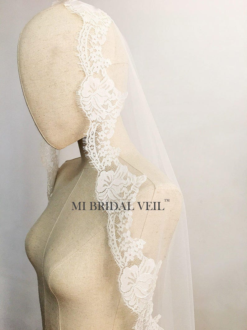 May include: A white bridal veil with a delicate floral lace trim. The lace is intricately designed with detailed flower patterns. The veil is draped over a mannequin, showcasing its flowing fabric and design. The text "MI BRIDAL VEIL" is visible.
