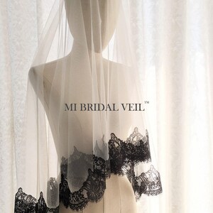 May include: A sheer, ivory-colored bridal veil with a delicate black lace trim. The veil is draped over a mannequin, showcasing the intricate floral pattern of the lace. The text "MI BRIDAL VEIL" is visible on the veil.
