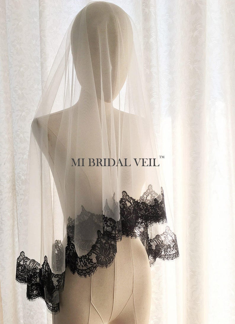 May include: A sheer, ivory-colored bridal veil with a delicate black lace trim. The veil is draped over a mannequin, showcasing the intricate floral pattern of the lace. The text "MI BRIDAL VEIL" is visible on the veil.