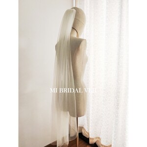 Può includere: Un velo da sposa color avorio trasparente è drappeggiato su un manichino. Il velo è lungo e fluente, con una consistenza morbida e delicata. Il testo "MI BRIDAL VIET" è visibile in basso. Lo sfondo è un muro bianco e una tenda a motivi.