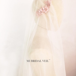 May include: A bridal veil with a delicate floral headpiece. The veil is a soft white, and the headpiece features pink flowers with sparkling accents. The text "MI BRIDAL VEIL" is visible on the veil.