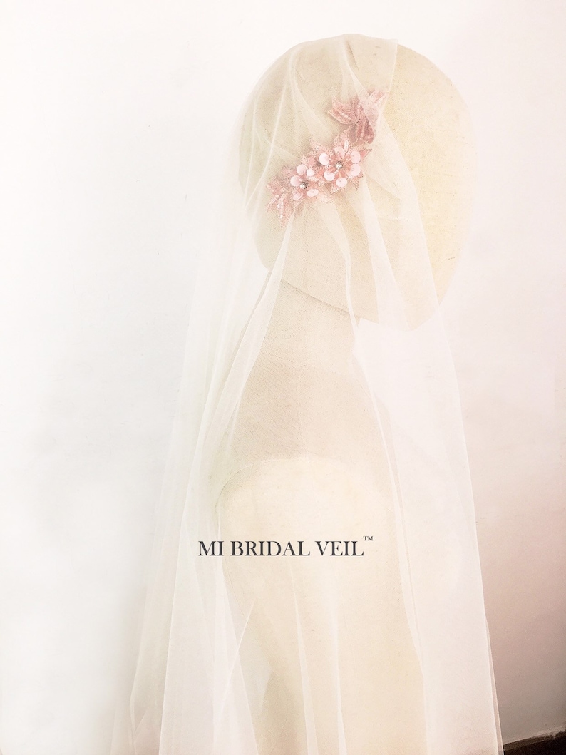 May include: A bridal veil with a delicate floral headpiece. The veil is a soft white, and the headpiece features pink flowers with sparkling accents. The text "MI BRIDAL VEIL" is visible on the veil.