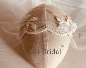 Puede incluir: Tocado de novia blanco roto con ribete de encaje y detalles florales. El tocado tiene hojas bordadas y pequeños adornos. El velo es de tela transparente, creando un aspecto suave y romántico. El texto "MI Bridal" es visible.