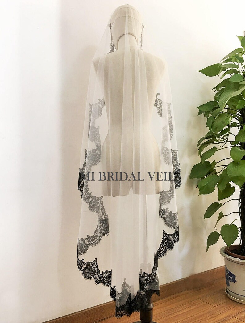 May include: A sheer white bridal veil with a delicate black lace trim and accents. The veil is displayed on a mannequin, showcasing its flowing design. The text "MI BRIDAL VEIL" is visible on the veil.