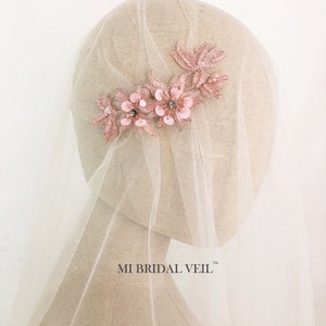 May include: A bridal veil with a floral headpiece. The headpiece is made of pink fabric flowers and leaves, embellished with sequins and small crystals. The veil is a sheer, off-white fabric. The text "MI BRIDAL VEIL" is visible.