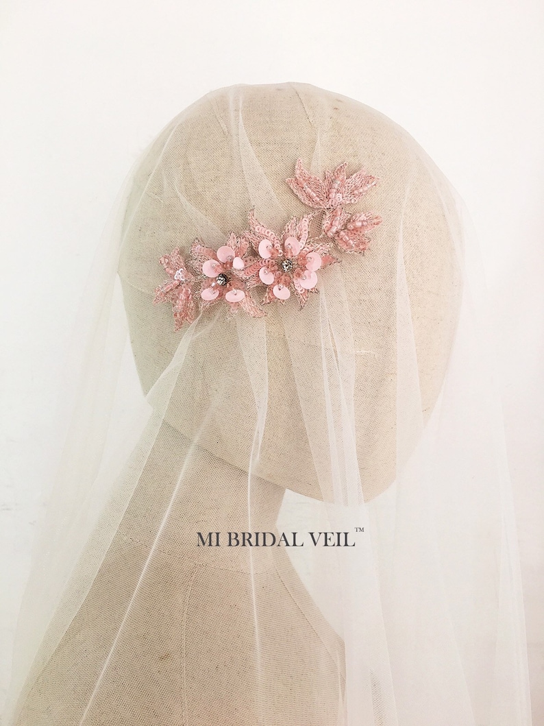 May include: A bridal veil with a floral headpiece. The headpiece is made of pink fabric flowers and leaves, embellished with sequins and small crystals. The veil is a sheer, off-white fabric. The text "MI BRIDAL VEIL" is visible.
