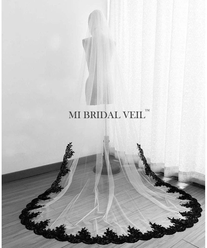 May include: A long, white bridal veil with a black lace trim, displayed on a mannequin. The veil is sheer, allowing light to pass through, and the black lace creates a decorative border along the edge. The text "MI BRIDAL VEIL" is visible.
