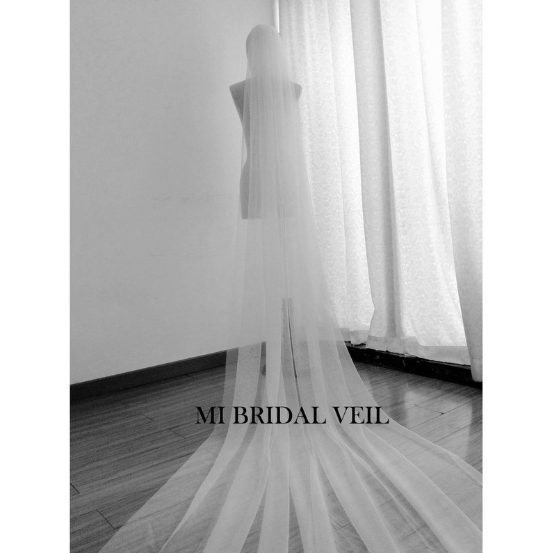 Pu&ograve; includere: Un lungo velo da sposa bianco e trasparente &egrave; drappeggiato su un manichino. Il velo scende fino al pavimento, creando un effetto fluido. Il testo "MI BRIDAL VEIL" &egrave; stampato in basso nell'immagine.