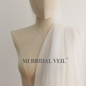 Pu&ograve; includere: Un velo da sposa bianco e trasparente drappeggiato su un manichino. Il velo &egrave; realizzato in un tessuto leggero e il testo "MI BRIDAL VEIL" &egrave; visibile. Lo sfondo &egrave; di colore bianco sporco.
