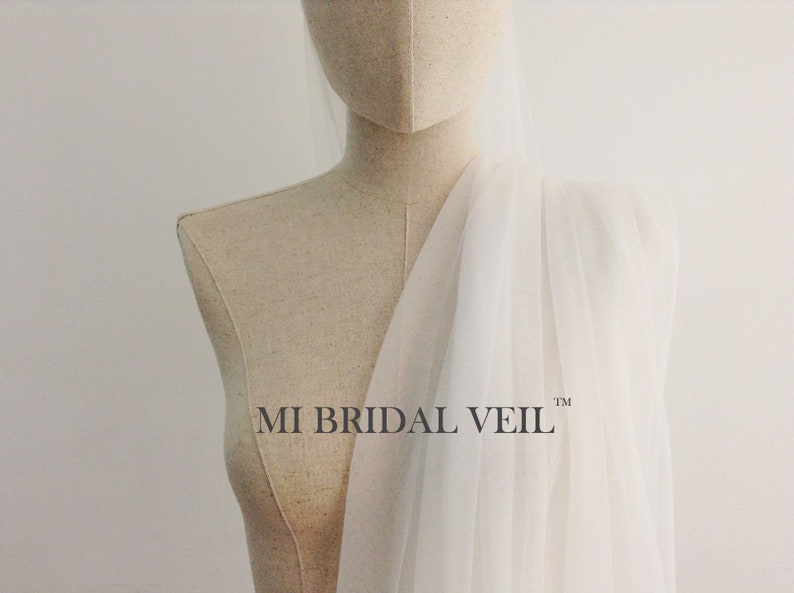 Pu&ograve; includere: Un velo da sposa bianco e trasparente drappeggiato su un manichino. Il velo &egrave; realizzato in un tessuto leggero e il testo "MI BRIDAL VEIL" &egrave; visibile. Lo sfondo &egrave; di colore bianco sporco.