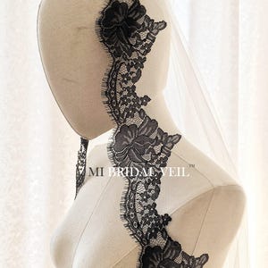 May include: A bridal veil with a delicate black lace trim featuring floral designs. The veil is sheer and attached to a mannequin head. The text "MI BRIDAL VEIL" is visible on the mannequin.