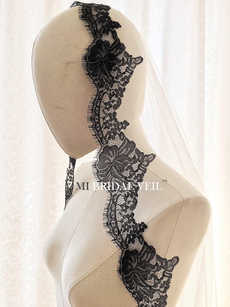 May include: A bridal veil with a delicate black lace trim featuring floral designs. The veil is sheer and attached to a mannequin head. The text "MI BRIDAL VEIL" is visible on the mannequin.