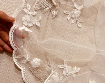 Puede incluir: Un delicado velo de novia blanco con bordado floral y borde de encaje festoneado. El velo presenta intrincadas flores y hojas bordadas, y un peine para una colocación segura en el cabello.