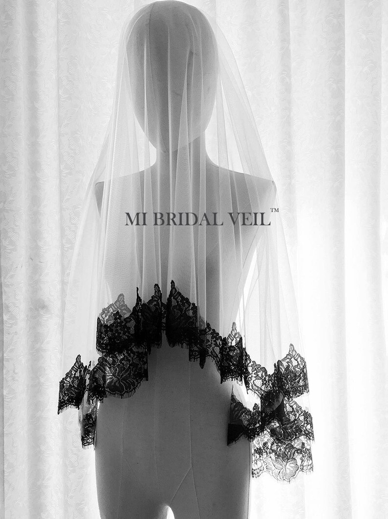 May include: A sheer white bridal veil with a black lace trim, displayed on a mannequin. The veil is long and flowing, with the text "MI BRIDAL VEIL" visible. The lace trim adds a decorative element to the veil.