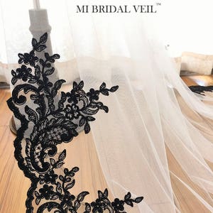 May include: A white bridal veil with a black floral lace trim. The lace features intricate designs of flowers and leaves. The text "MI BRIDAL VEIL" is visible on the veil.