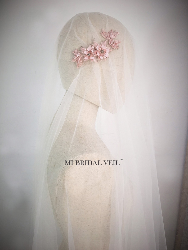 May include: A bridal veil with a delicate floral headpiece. The headpiece features pink fabric flowers and embellishments. The veil is sheer and white, draped over a mannequin. The text "MI BRIDAL VEIL" is visible.