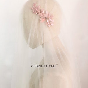 May include: A bridal veil with a delicate, light pink floral hairpiece. The hairpiece features leaves and flowers with sparkling accents. The veil is sheer and white, creating a soft, romantic look. The text "MI BRIDAL VEIL" is visible.