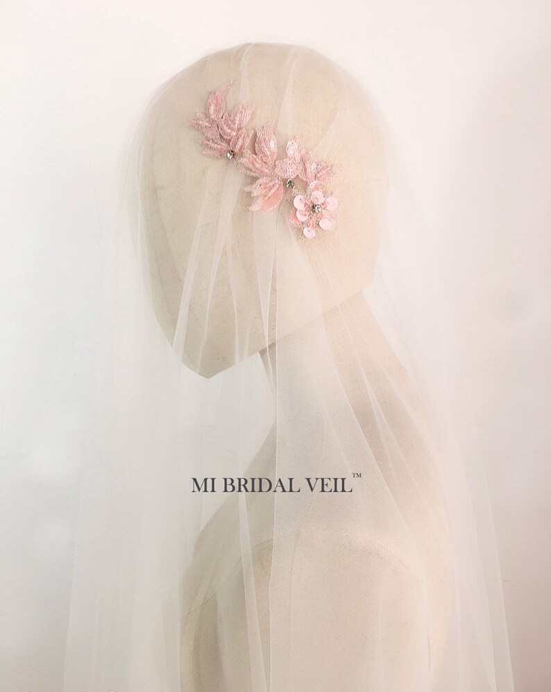 May include: A bridal veil with a delicate, light pink floral hairpiece. The hairpiece features leaves and flowers with sparkling accents. The veil is sheer and white, creating a soft, romantic look. The text "MI BRIDAL VEIL" is visible.
